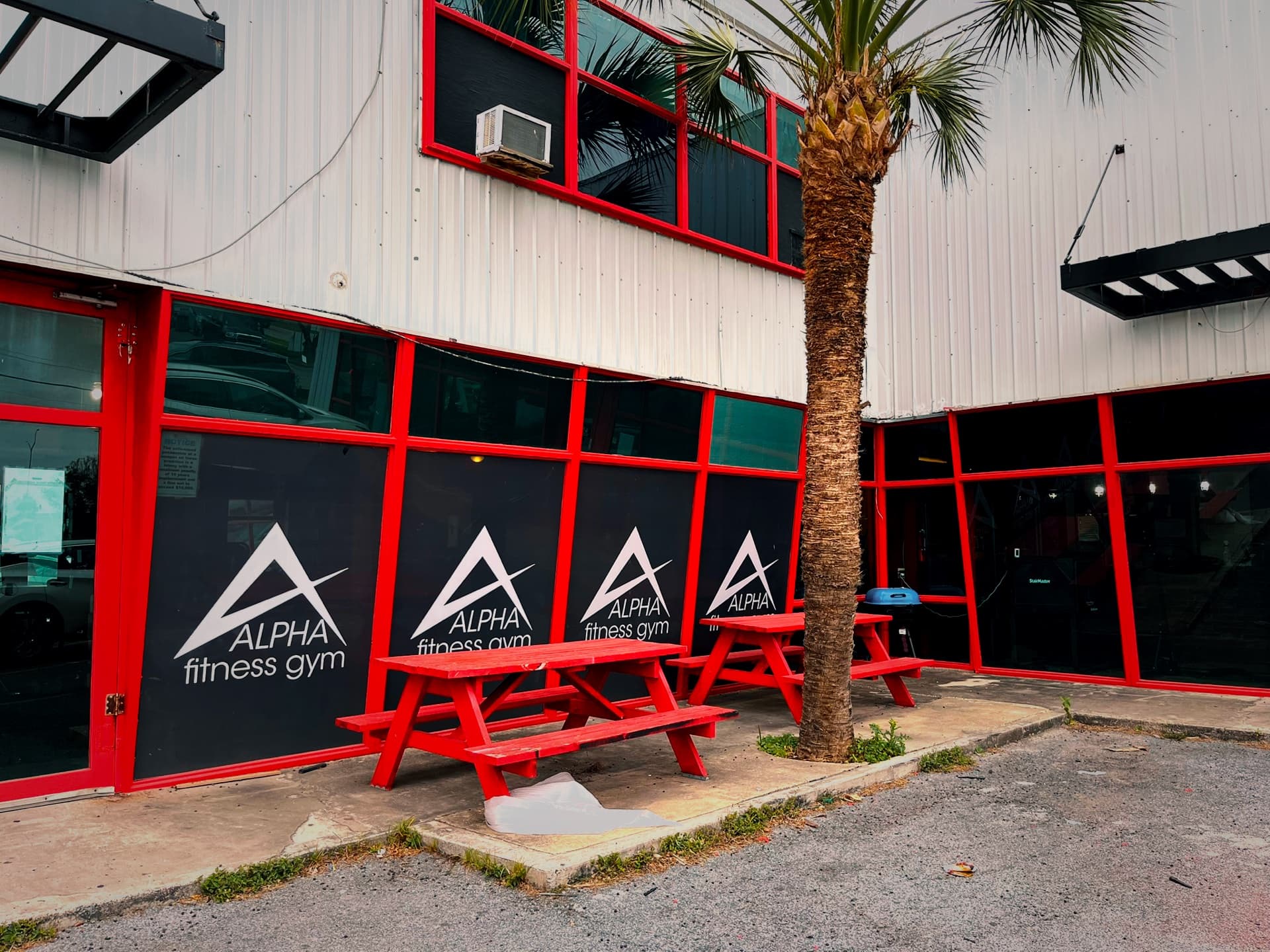 The Alpha Fitness building exterior, black windows framed in red, with a red picnic table and a palm tree out front